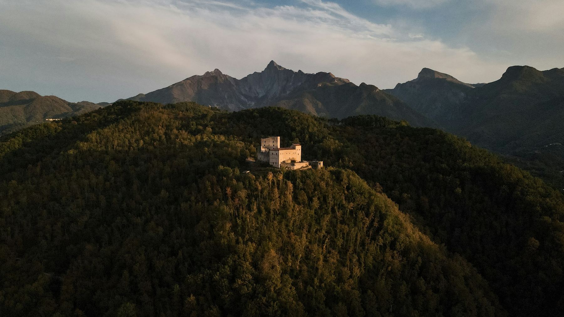 Lunigiana: eine Burg über bewaldeten Hängen, dahinter die Apuanischen Alpen